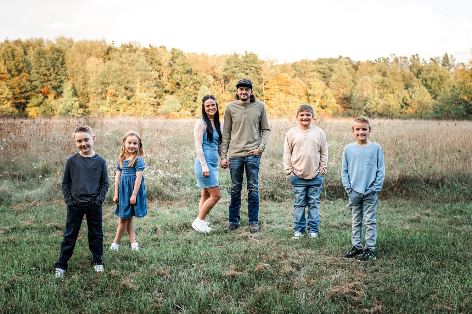 Family of six smiling in an open field with autumn trees in the background on a sunny day.