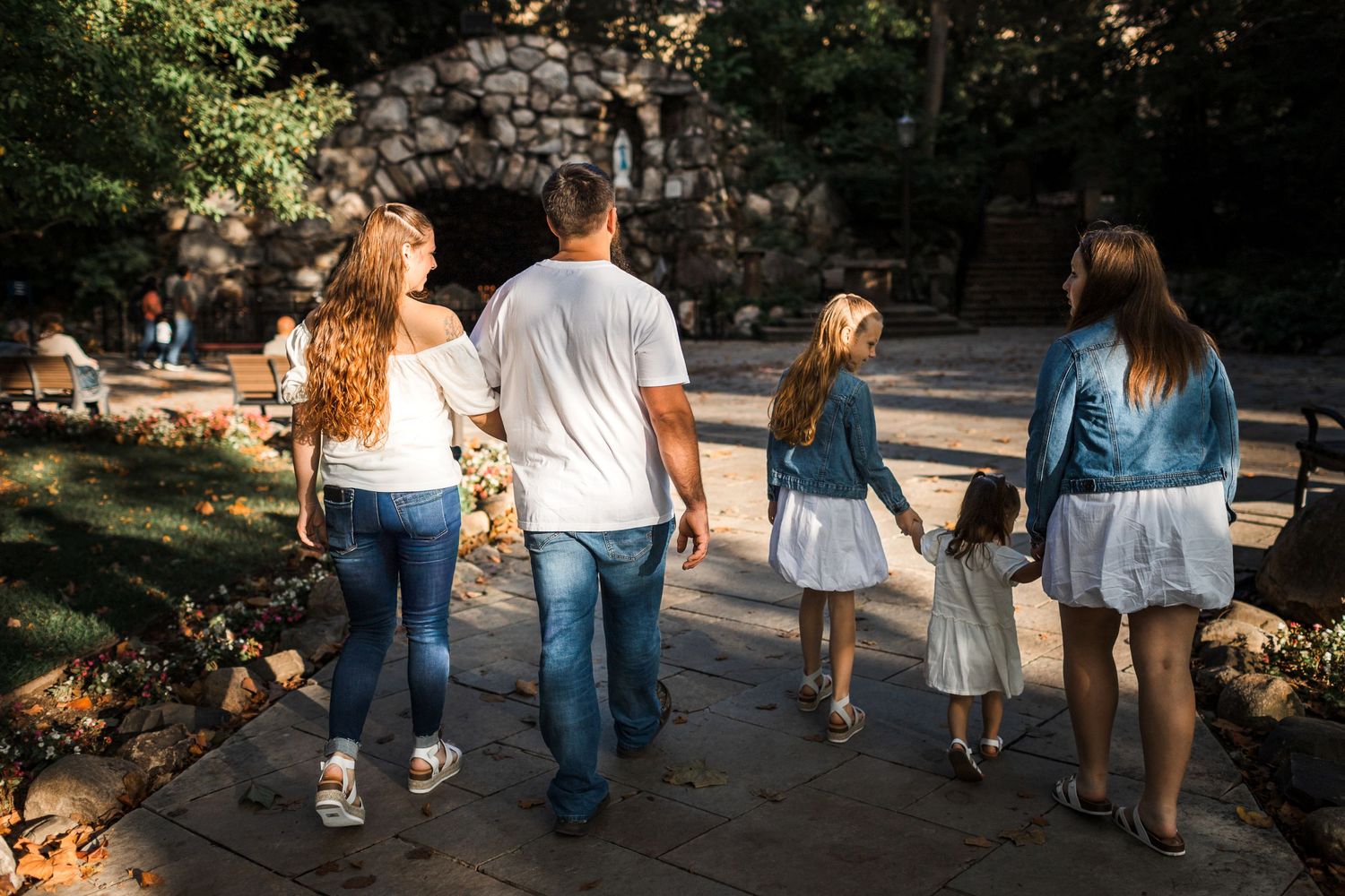Family of five walking together on a stone path through a sunny park, viewed from behind.