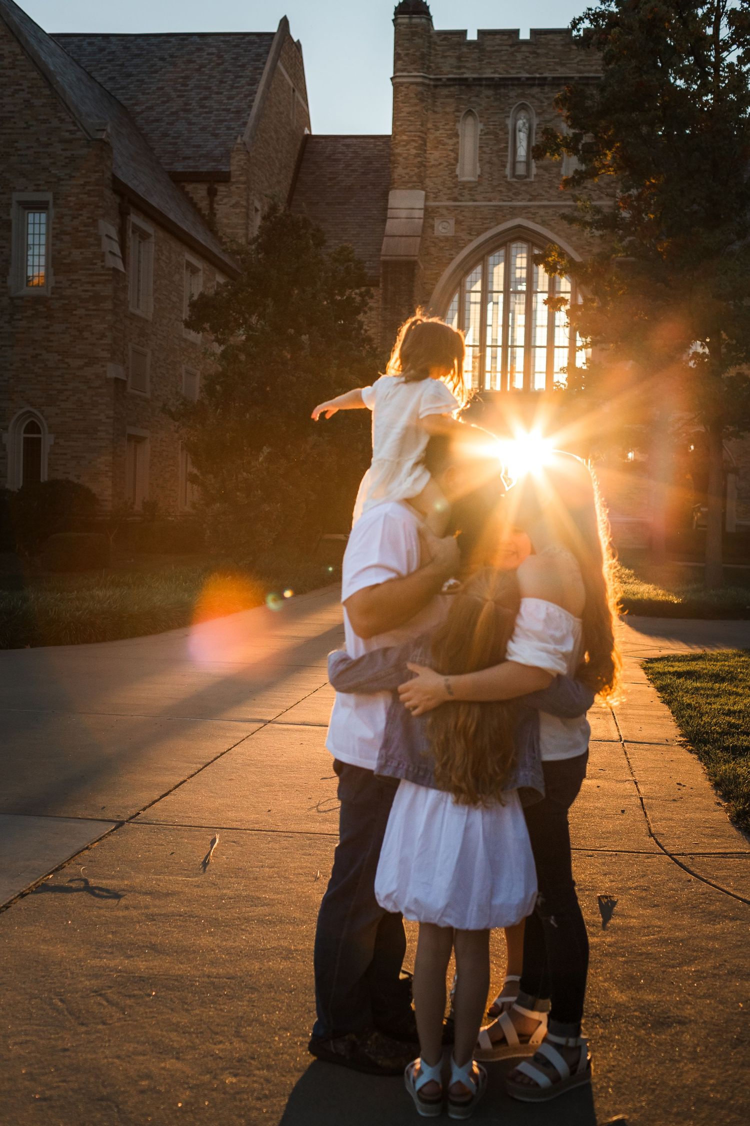 Family embracing at sunset in front of a stone church, with golden lens flare and a child perched on shoulders.