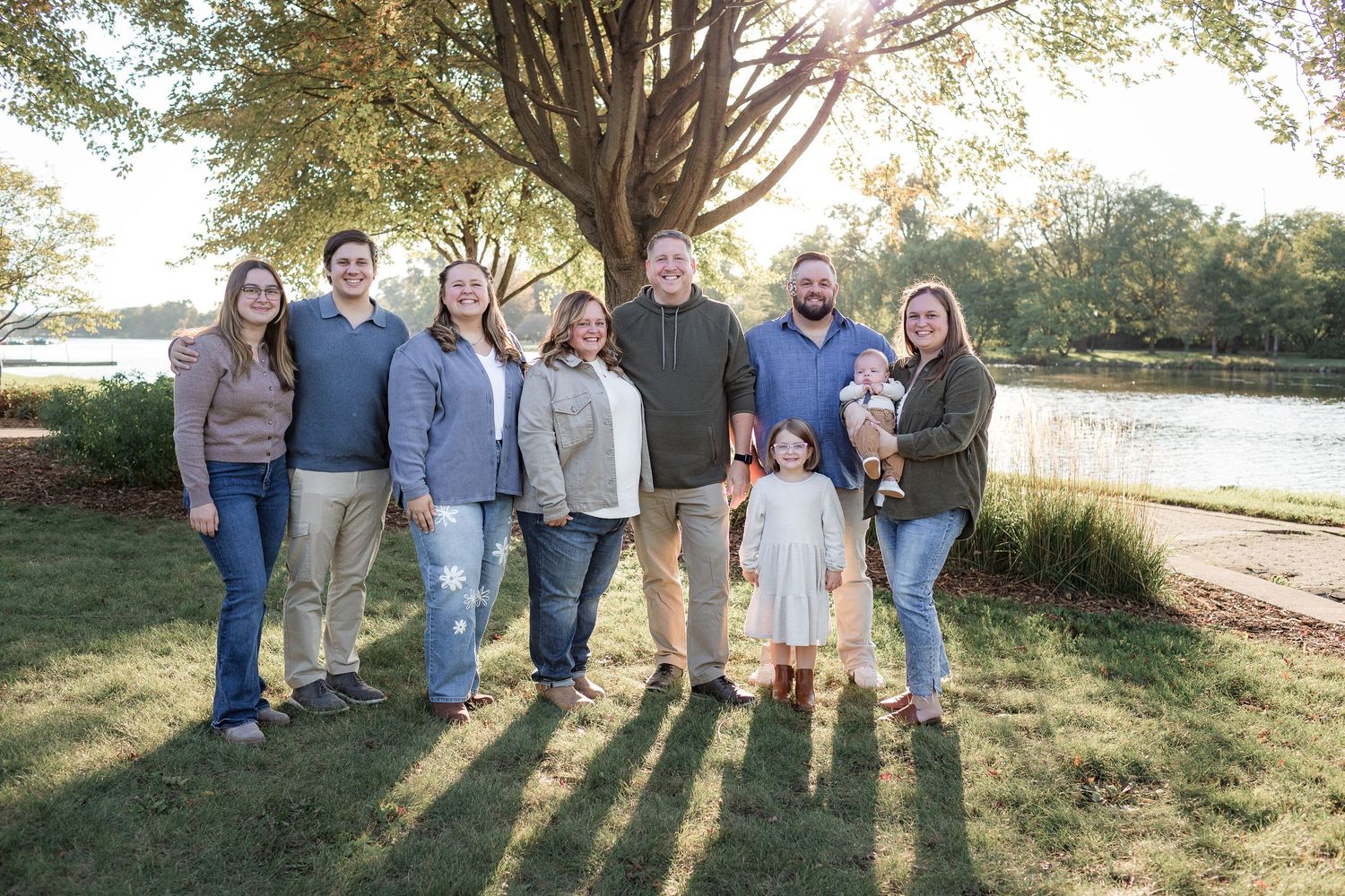 Family of eight posing outdoors near a serene lake surrounded by lush green trees on a sunny day.