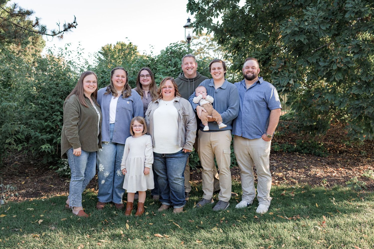 Family of eight posing outdoors on green grass with lush trees, including adults, children, and a baby.