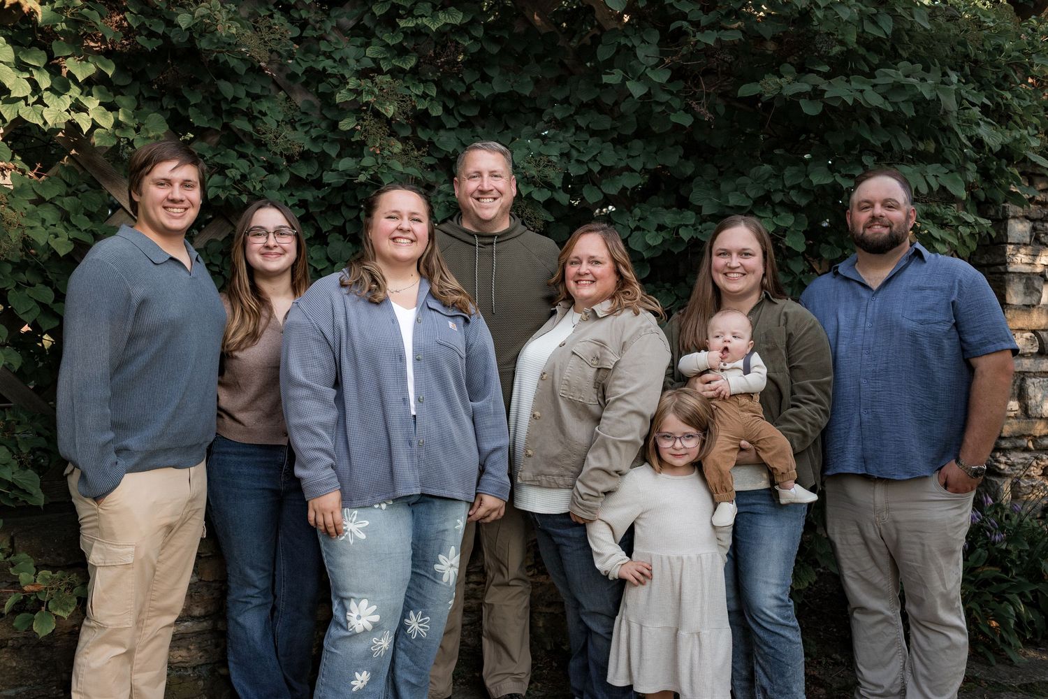 Large family group portrait smiling outdoors against stone wall with lush green ivy backdrop.