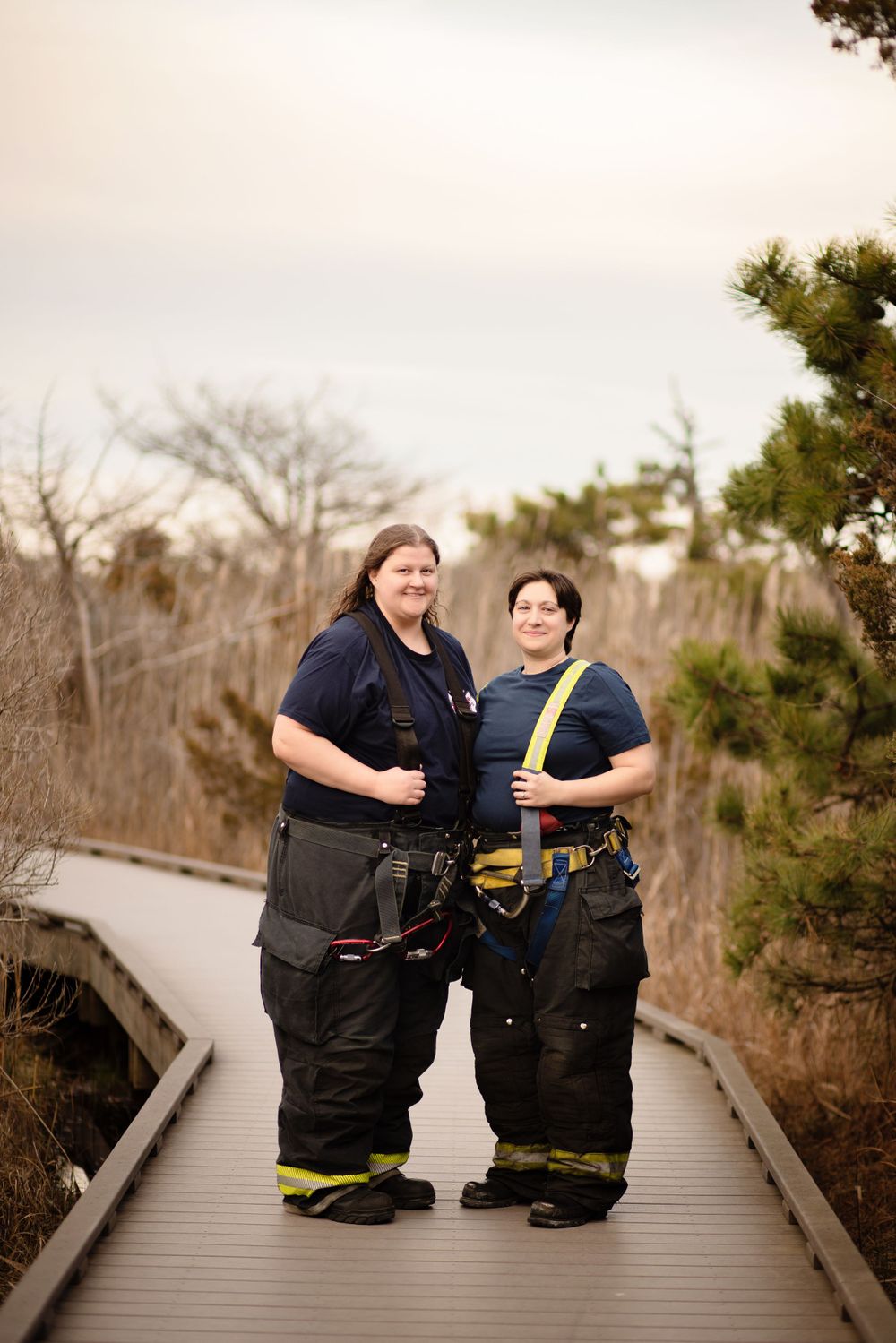 Two firefighters in full turnout gear stand smiling together on a wooden boardwalk surrounded by winter brush.