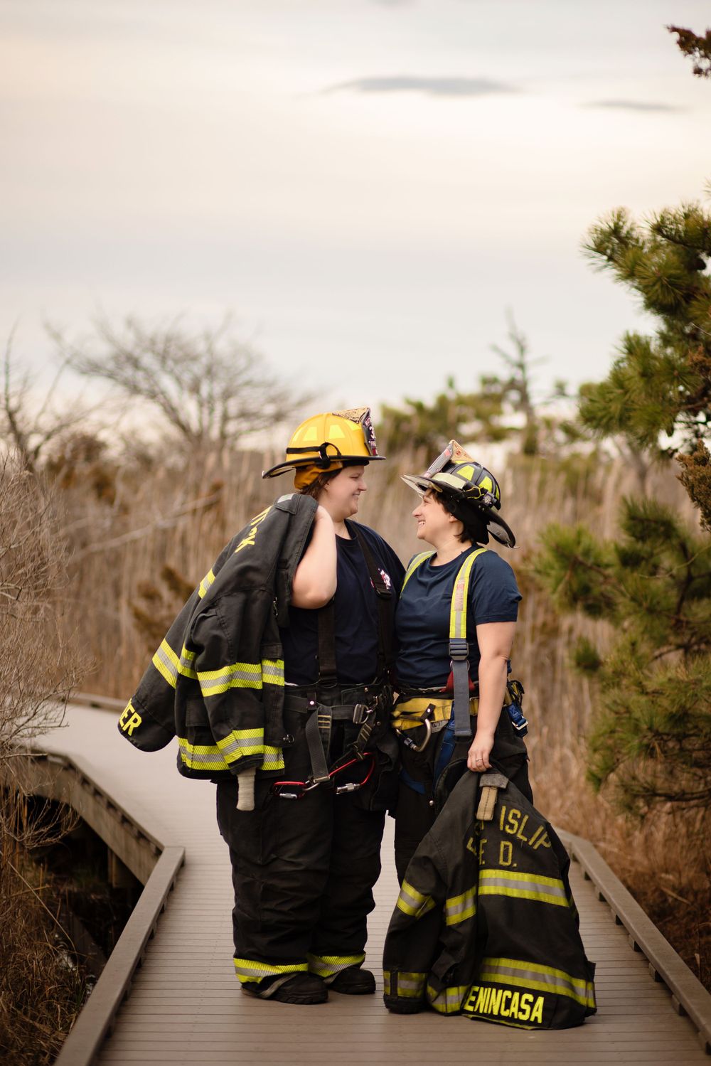 Two firefighters in full protective gear stand together on a wooden boardwalk surrounded by nature.