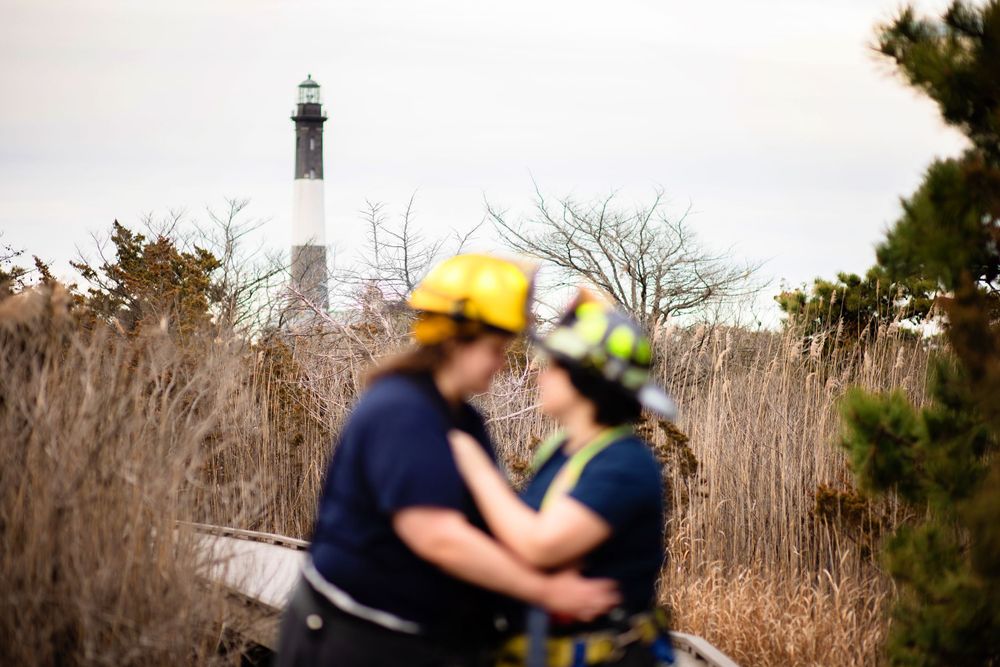 A lighthouse stands tall in the background while two people in fire helmets embrace among tall grass and foliage.