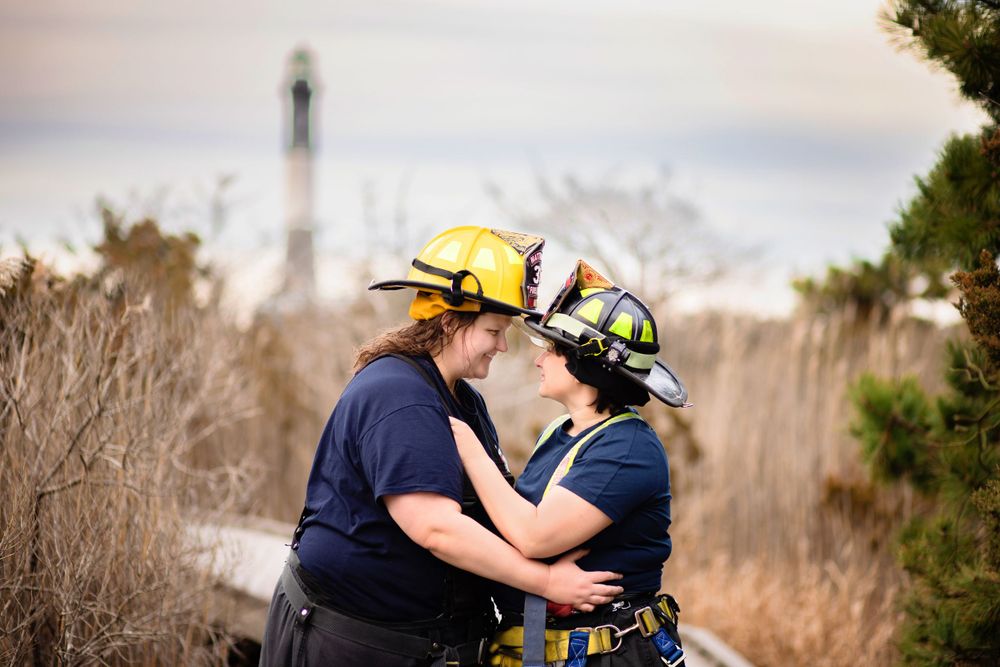 Two firefighters in protective gear embrace in front of tall dry grass with a lighthouse visible in the background.