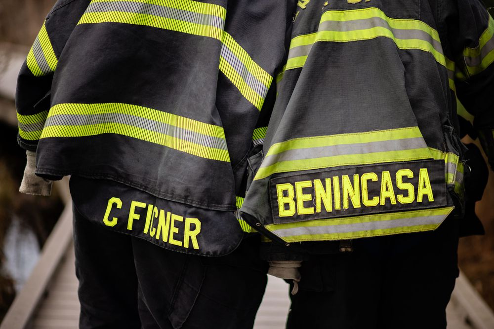 Close-up of firefighter turnout gear showing reflective stripes and name tags reading C FINER and BENINCASA.
