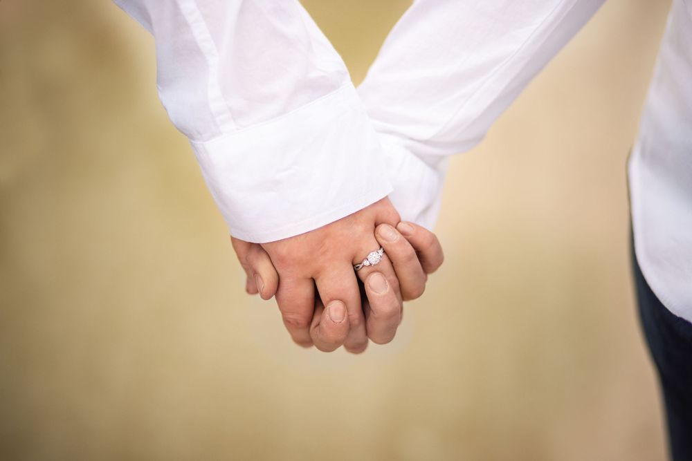 Two hands tenderly clasped together while wearing white clothing against a beige background.
