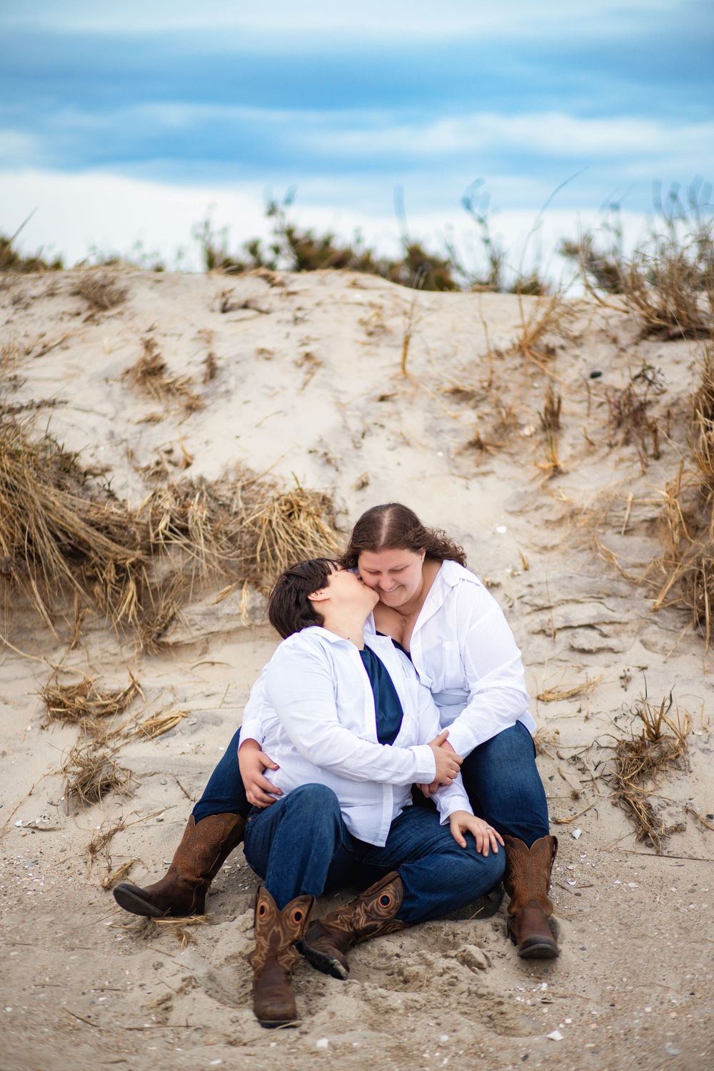 A couple shares a tender moment while sitting together on sandy dunes against a blue cloudy sky.