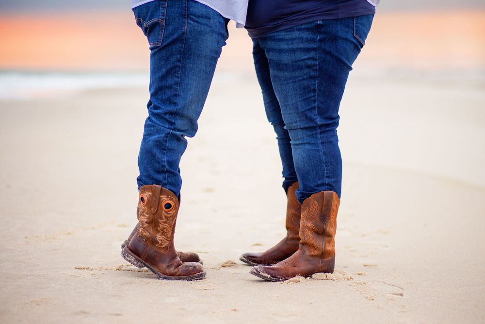 Person in blue jeans and brown cowboy boots standing on a sandy beach at sunset.