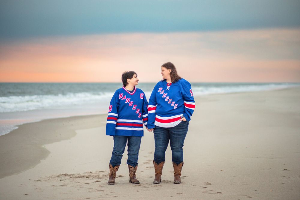 Two people in New York Rangers hockey jerseys walking together on a beach at sunset with matching outfits.