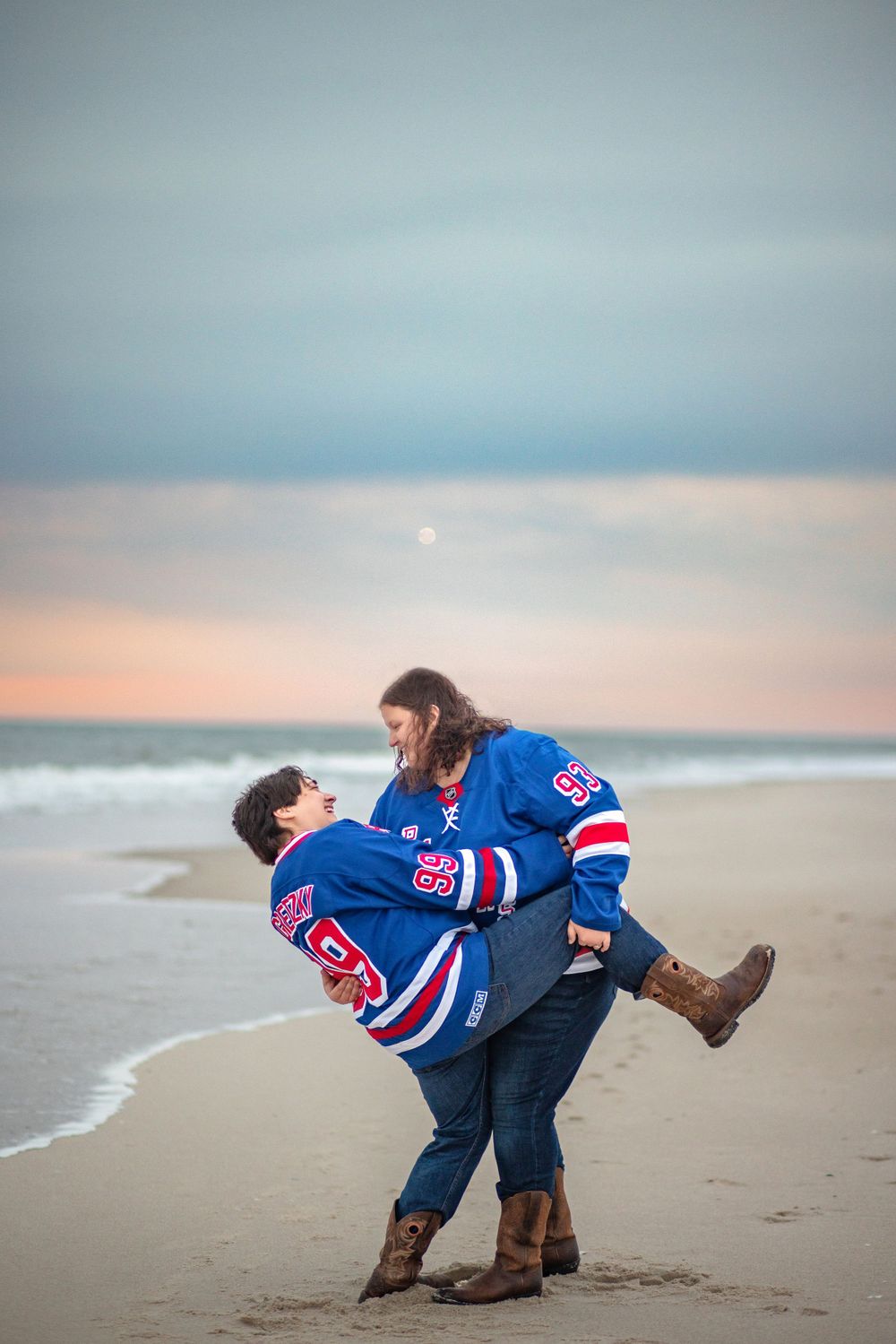 Two people wearing matching New York Rangers hockey jerseys share a romantic moment on the beach at sunset.