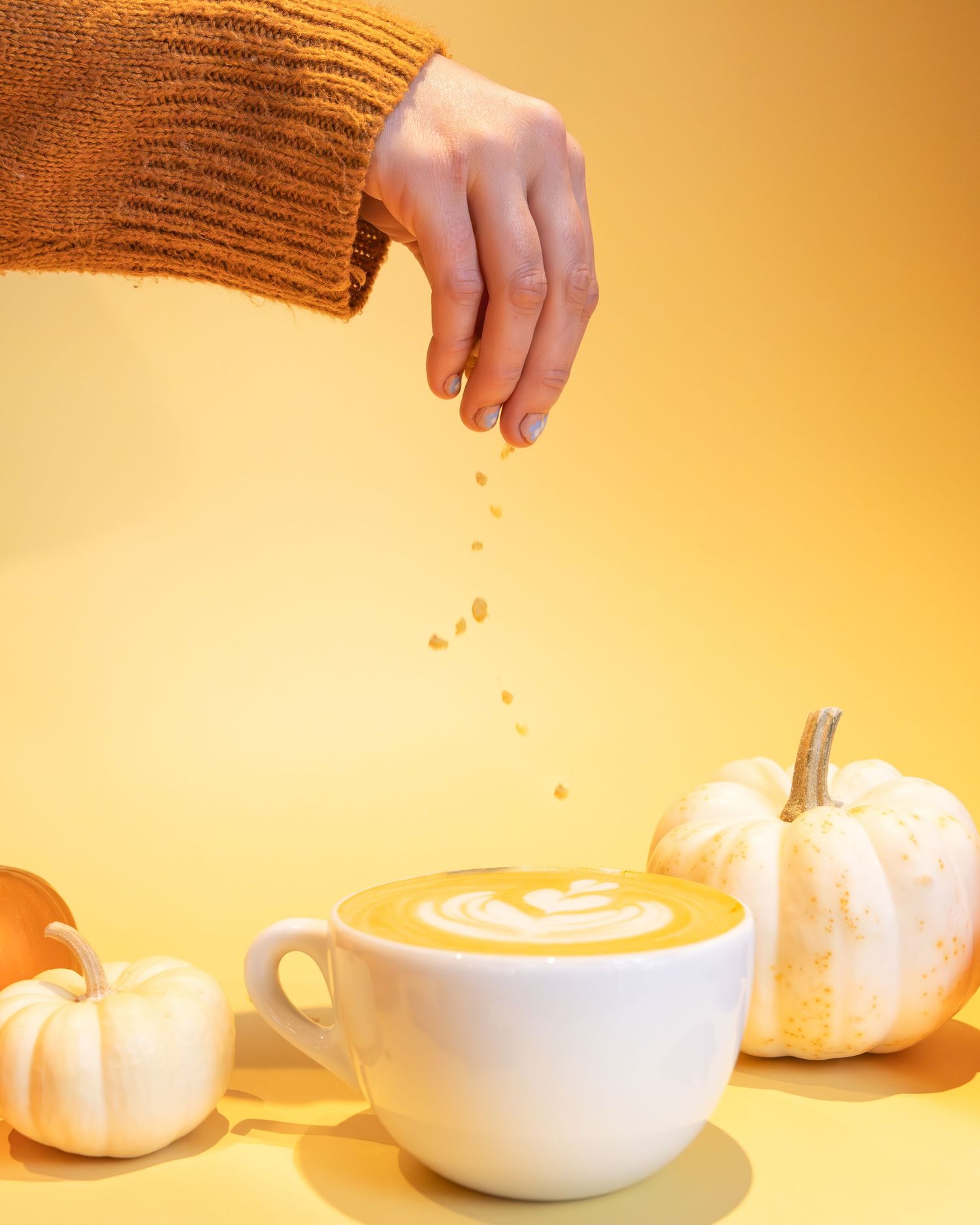 Hand in orange sweater sprinkles cinnamon over pumpkin spice latte with white pumpkin decorations on yellow background.