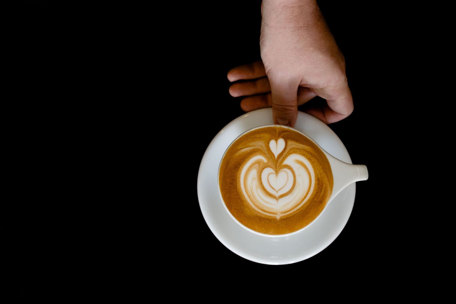 A hand holds a white coffee cup with intricate latte art design featuring a heart pattern in the foam.
