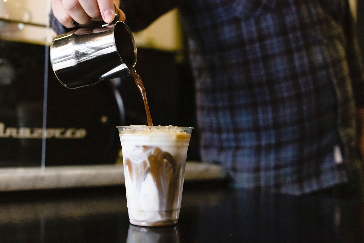 Coffee being poured from a metal pitcher into a glass filled with milk creating a layered iced drink effect.