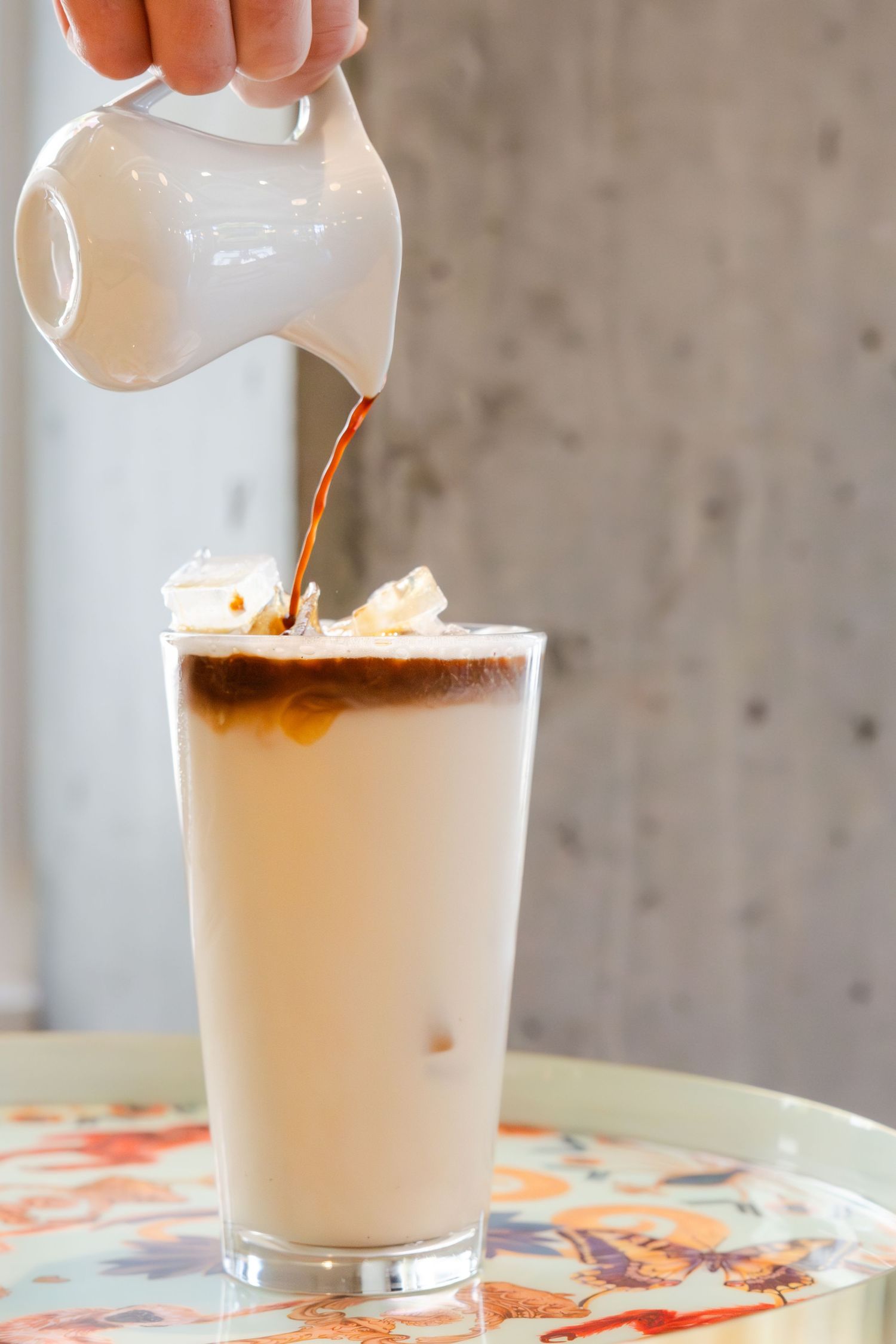 Creamy milk being poured into a tall glass of iced coffee on a decorative floral tray.