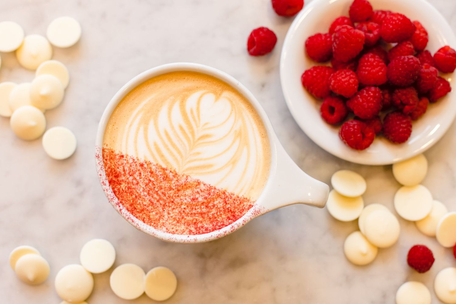 A pink latte with latte art sits alongside a plate of fresh raspberries and scattered white chocolate chips on marble.