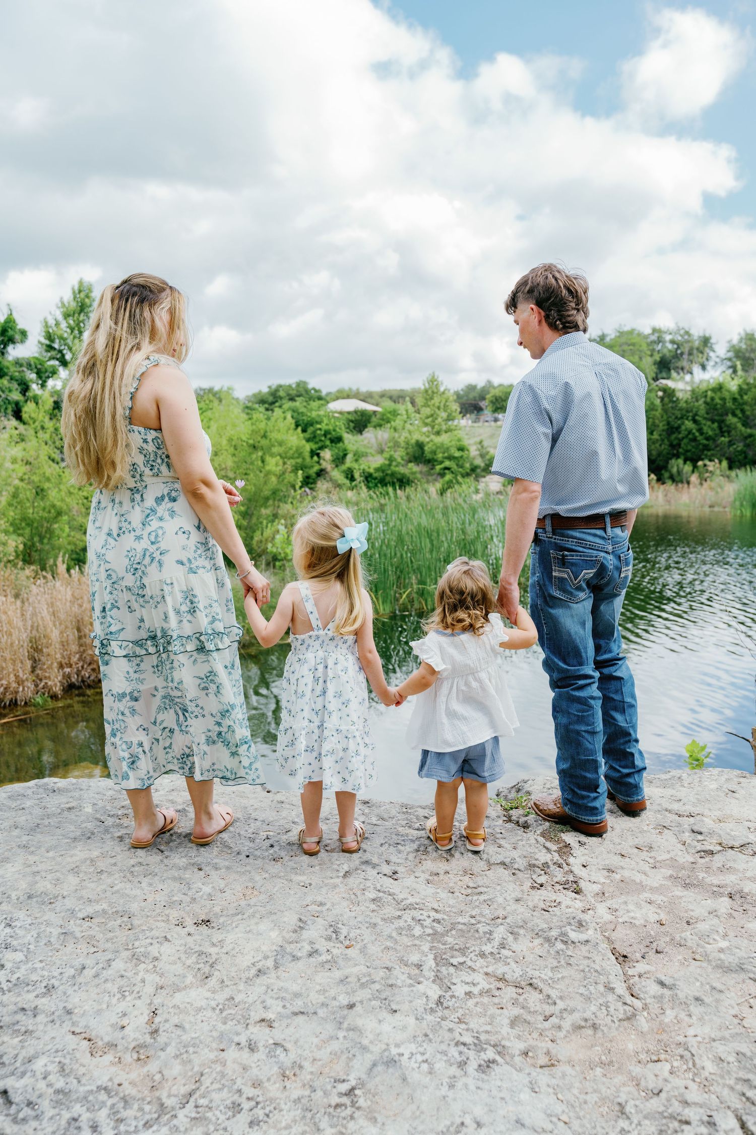 Family of four holding hands, viewed from behind, standing on rocks overlooking a serene pond surrounded by lush greenery.