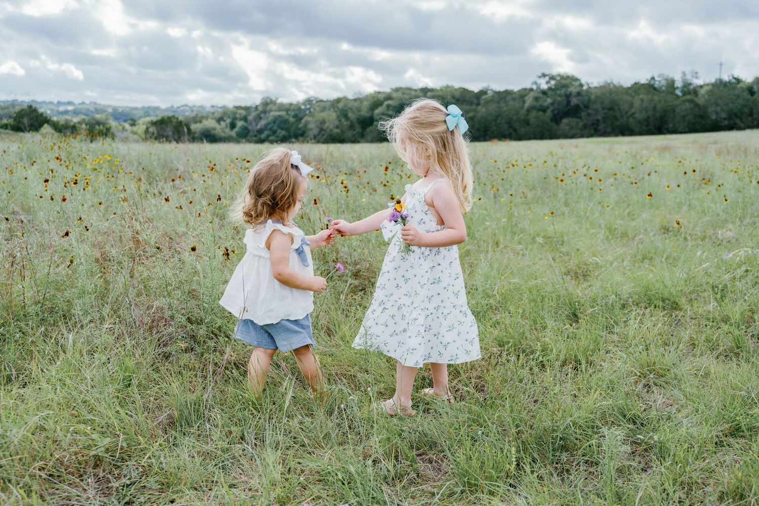Two young girls exchange wildflowers in a lush green meadow on a cloudy day.