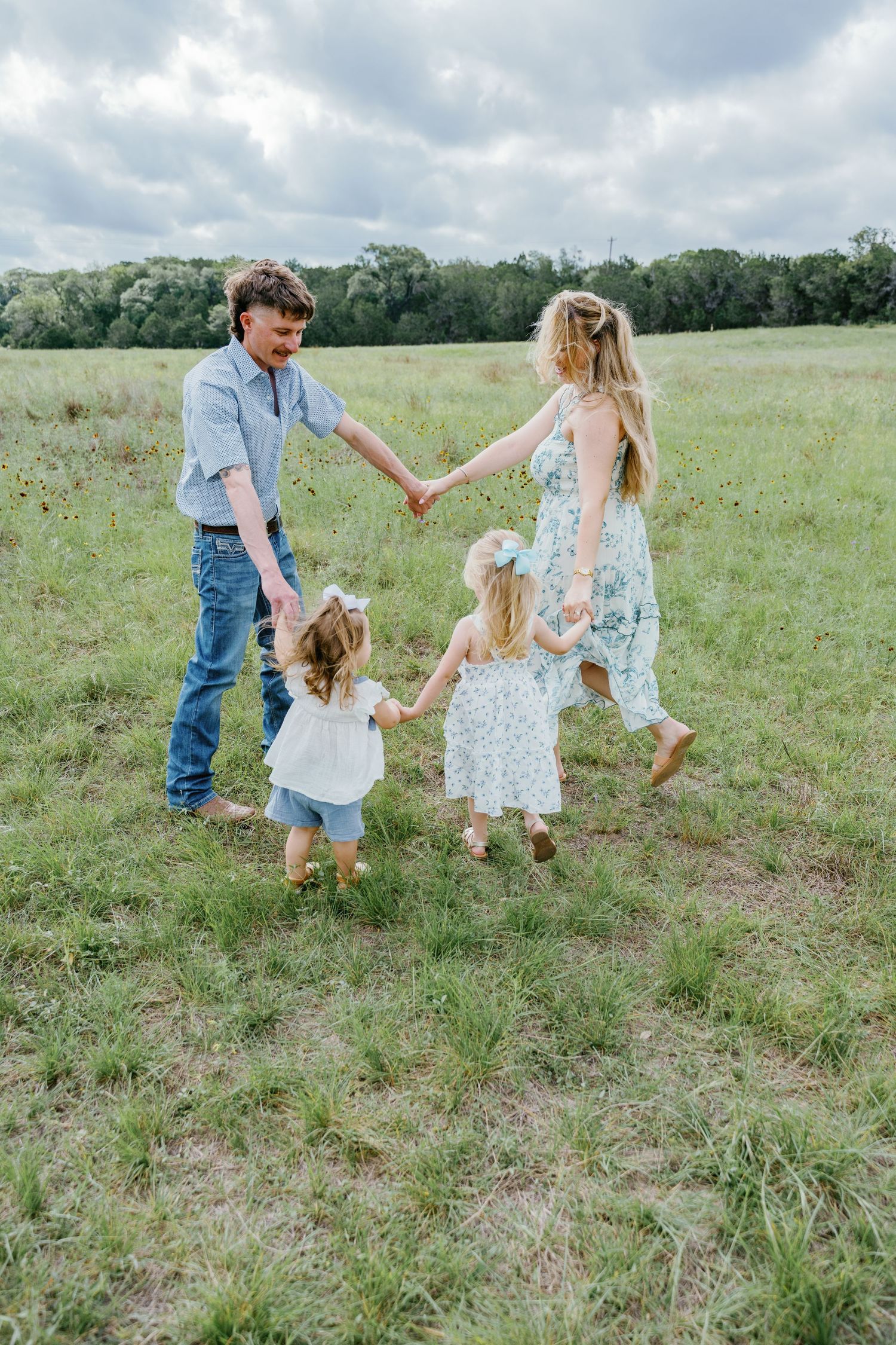 Family of four holding hands in a circle on a green meadow, with two young girls in white and floral dresses.