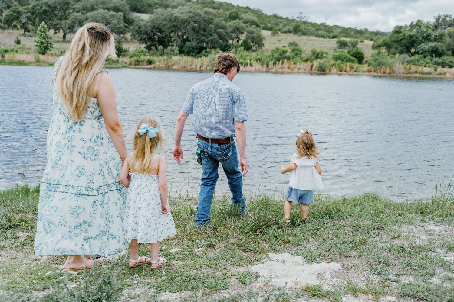 Family of four holding hands by a serene lake, two young girls in white dresses with parents on a grassy shore.