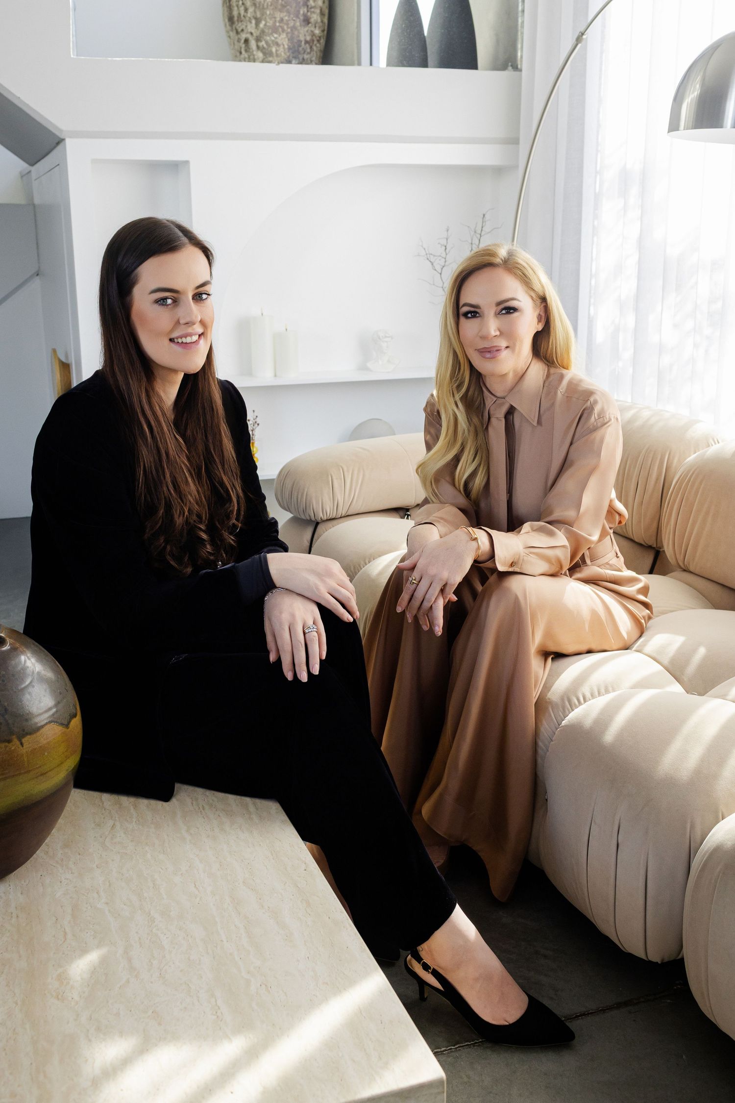 Two people in contrasting black and beige outfits sit casually on a light-colored couch in a bright modern space.