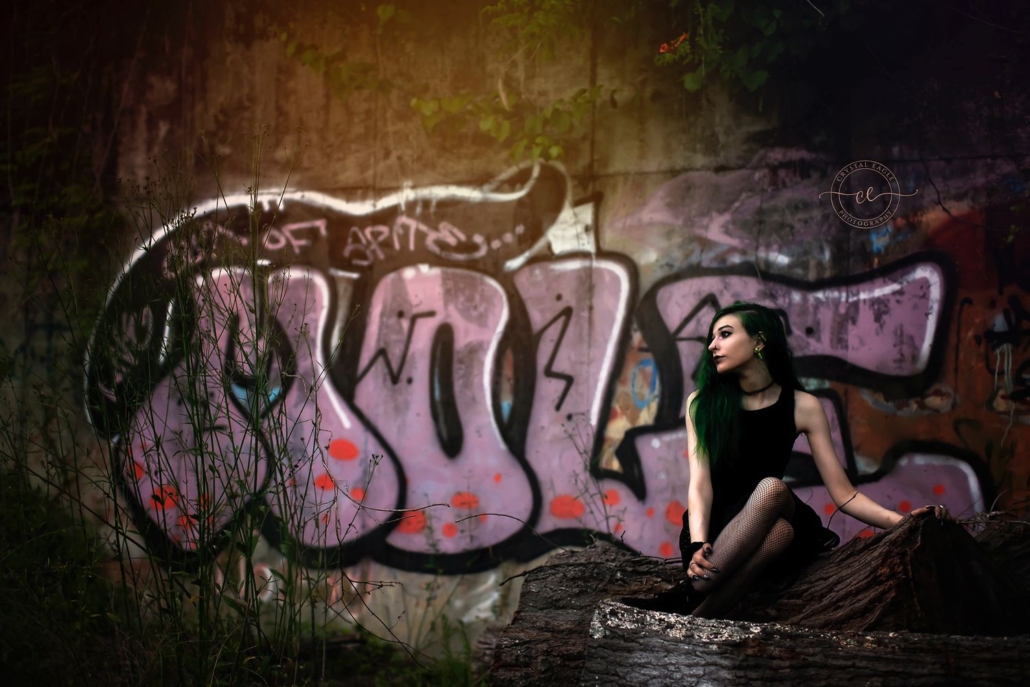 A moody urban portrait of a woman with green hair sitting by bold graffiti, blending edge, art, and quiet reflection.