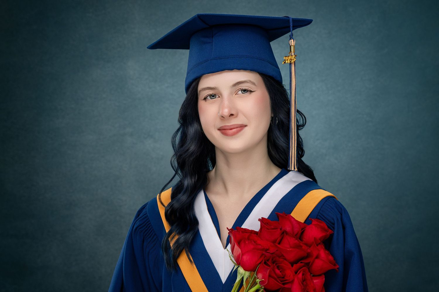 Graduate in bright blue cap and gown holds diploma while posing for graduation photos against dark background.