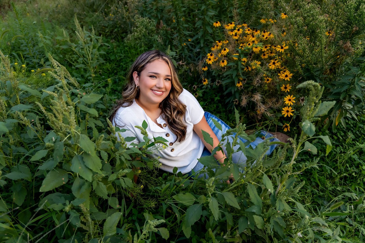 A person in white poses among vibrant yellow wildflowers and lush greenery in a garden setting.