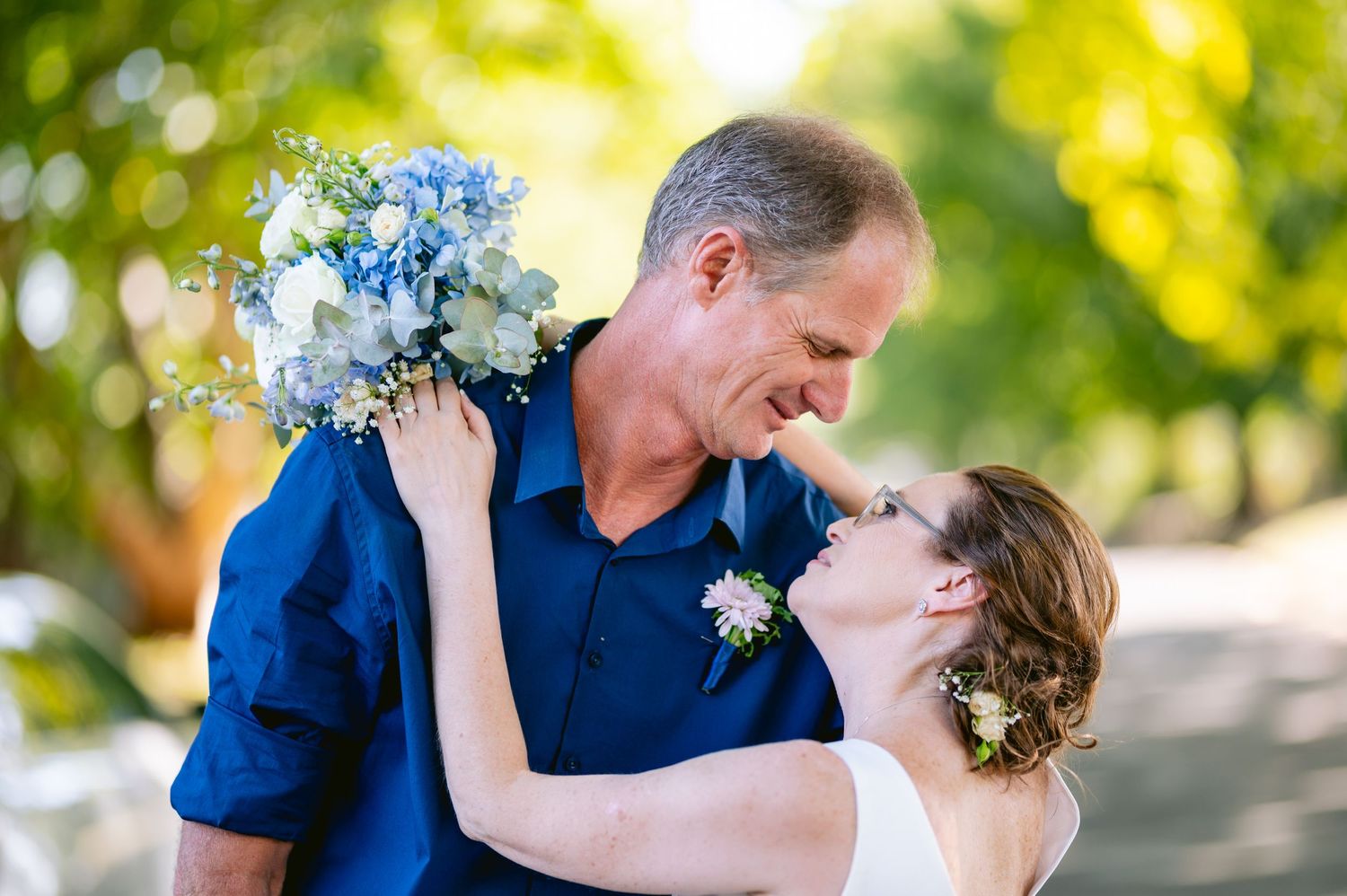 A couple in navy blue and white attire share romantic moments in a sun-dappled outdoor setting during their wedding.