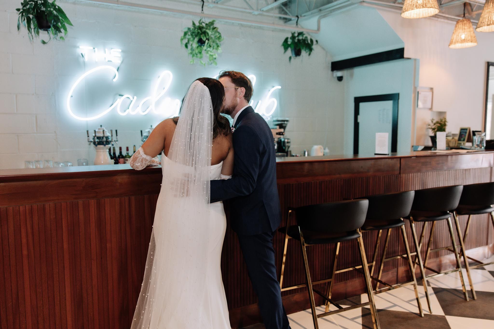 Newlyweds share an intimate moment at a bar with a glowing neon cocktail sign and wooden counter.