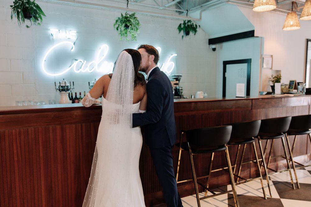 Newlyweds share an intimate moment at a bar with a glowing neon cocktail sign and wooden counter.