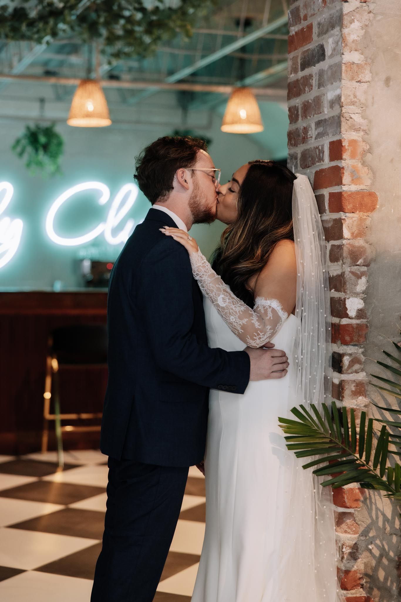 Newlyweds share intimate kisses against exposed brick wall in an industrial-chic venue with neon lighting.