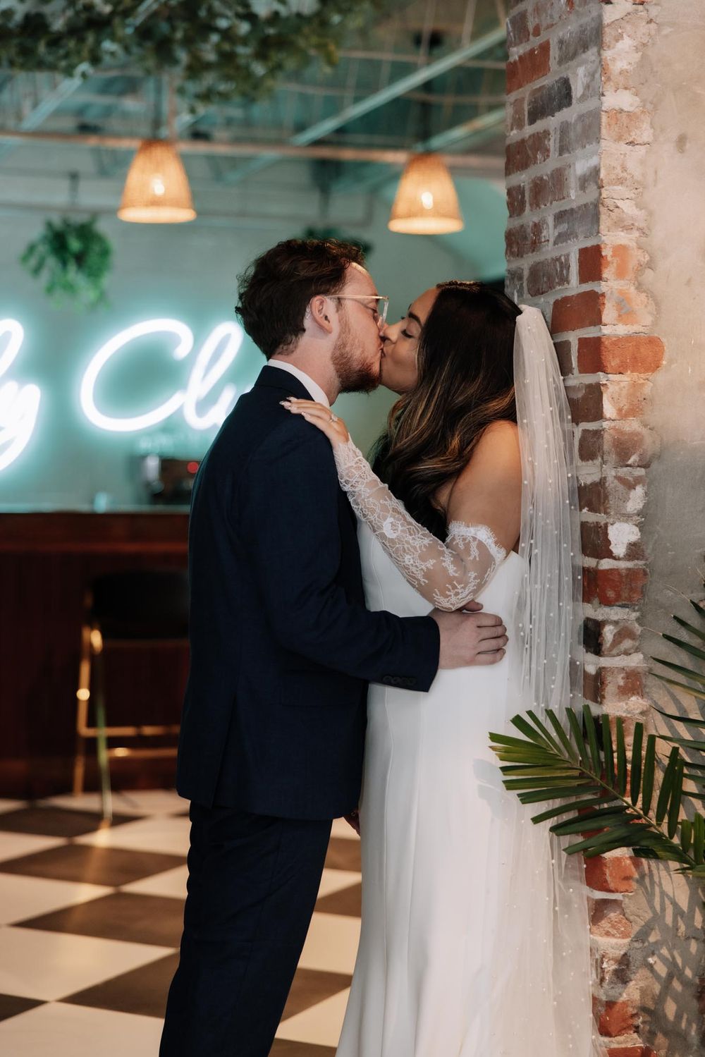Newlyweds share intimate kisses against exposed brick wall in an industrial-chic venue with neon lighting.