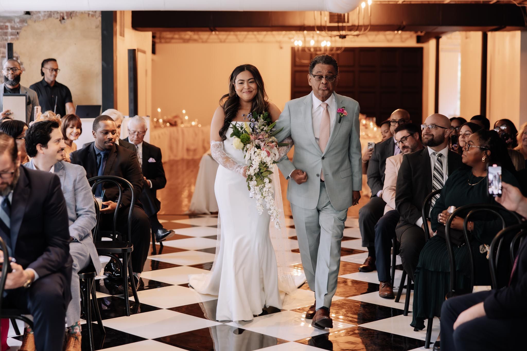 Wedding ceremony guests seated in rows watch the bride walk down the aisle in a warmly lit indoor venue.