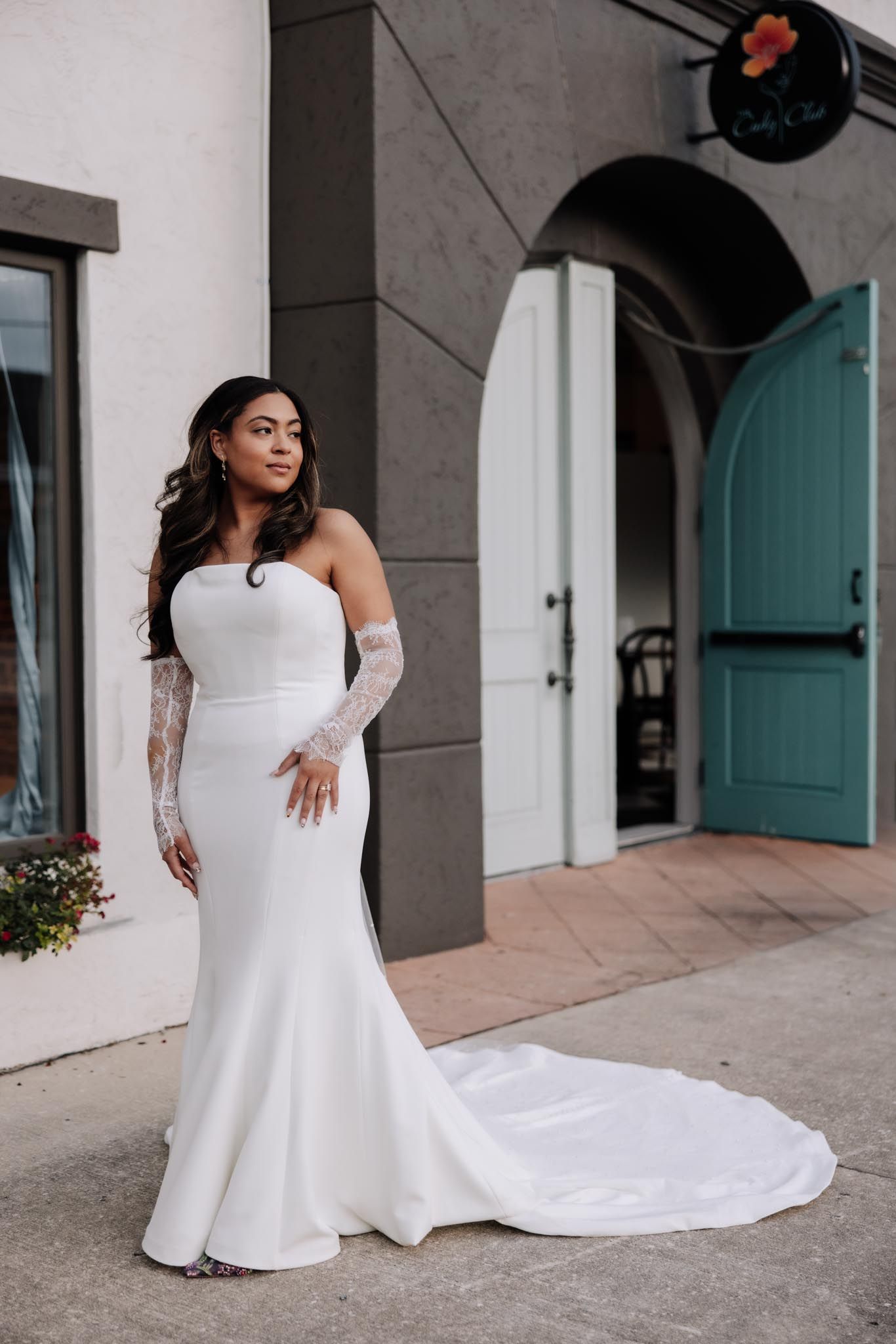A bride in a sleek white wedding gown with a flowing train poses on a porch with teal doors and columns.