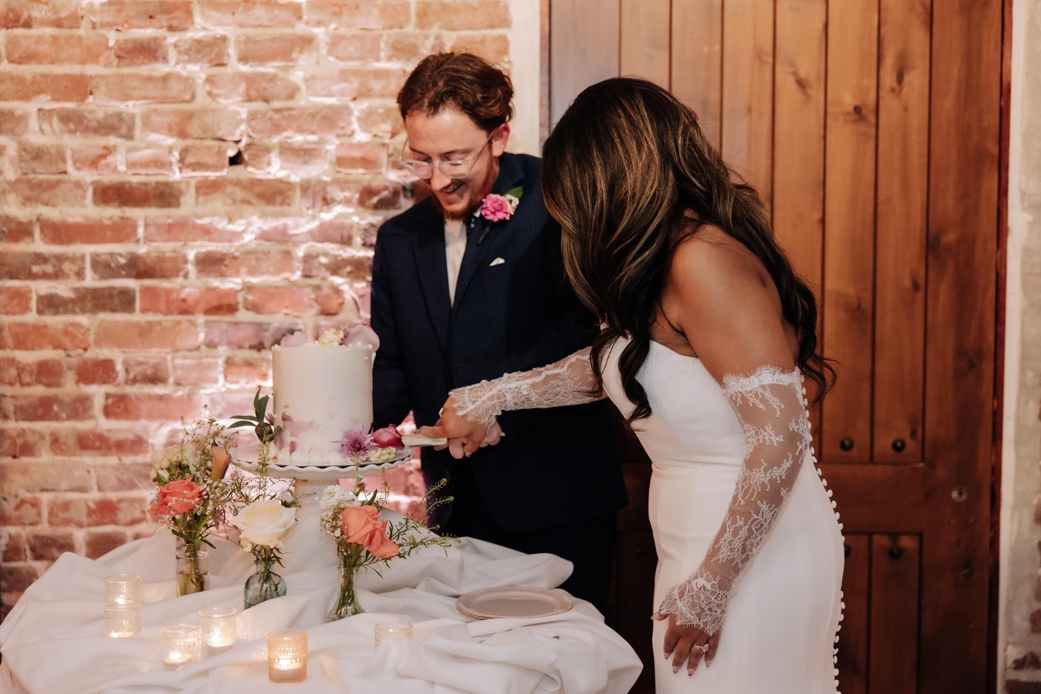A couple cuts into their elegant wedding cake together at a rustic brick-walled venue.