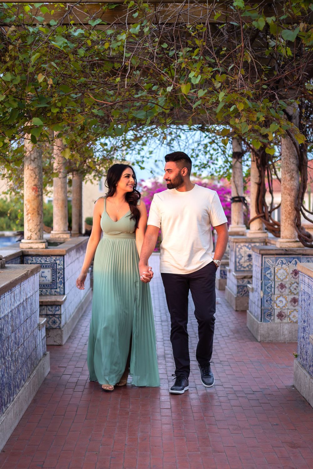 A couple walks hand in hand along a scenic pathway with decorative tile walls and lush green foliage overhead.