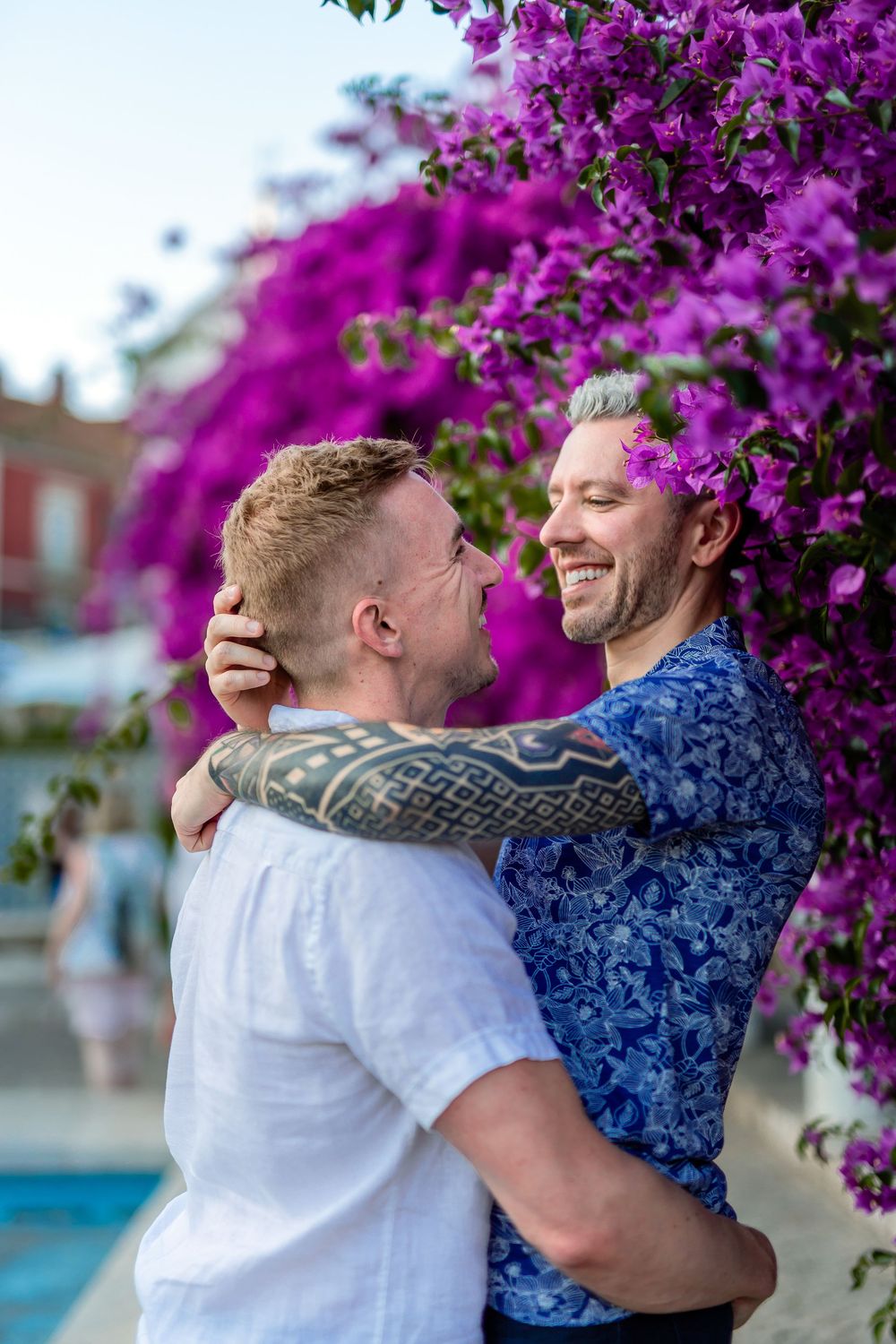 Two people embrace and laugh together under vibrant purple bougainvillea flowers on a sunny day.