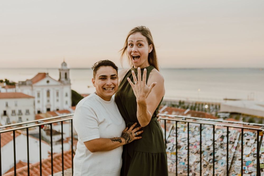 Couple embraces on a balcony overlooking a coastal town at sunset with ocean views in the background.