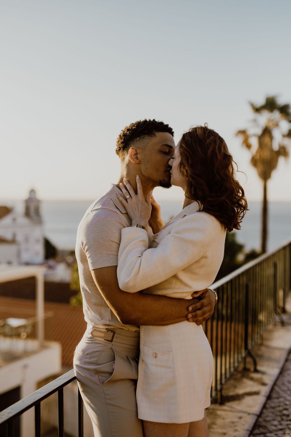 A romantic couple embraces on a balcony at sunset with palm trees and Spanish architecture in the background.
