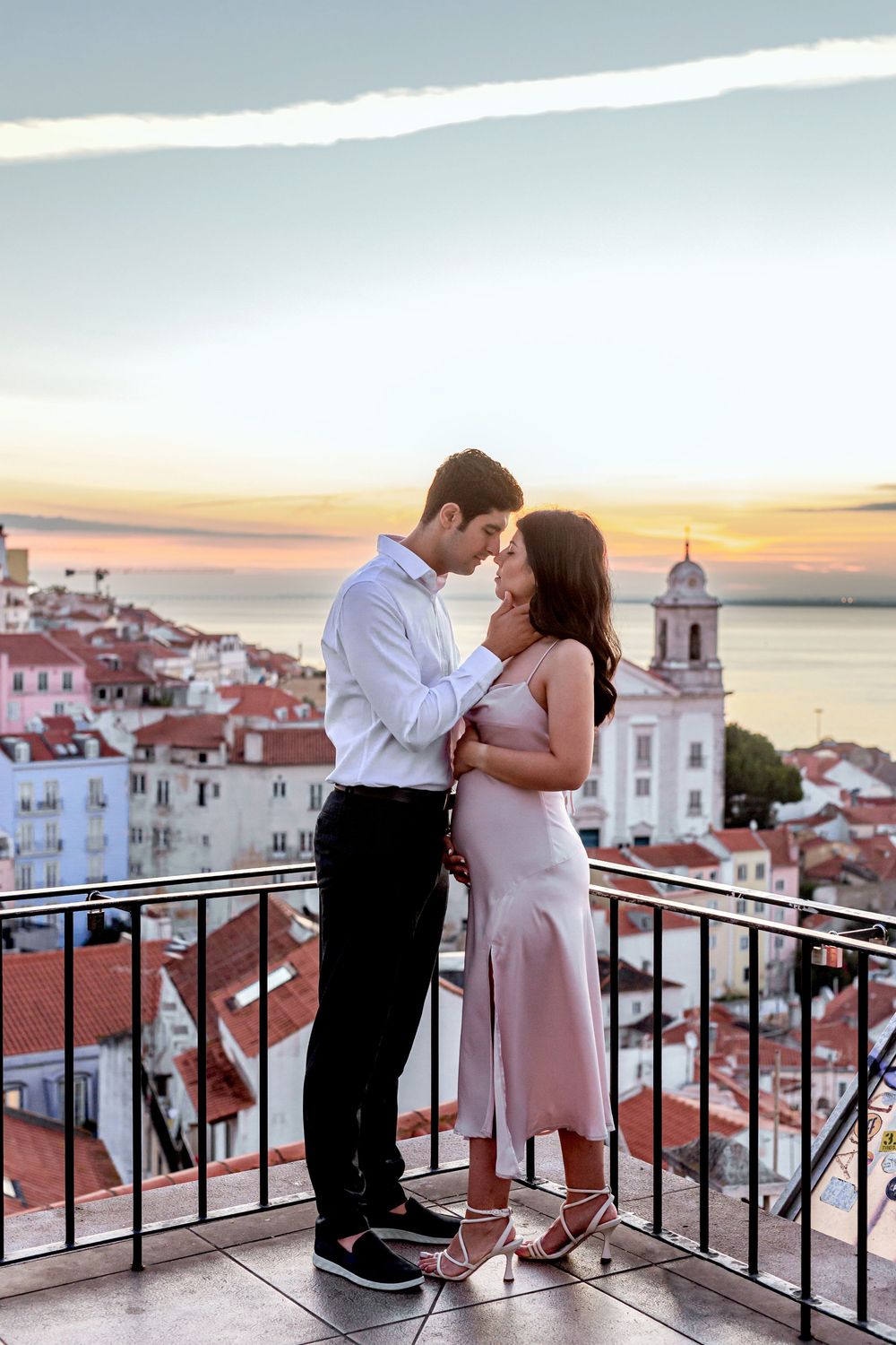 Romantic couple embracing on a balcony overlooking Lisbon's historic Alfama district with orange rooftops at sunset.