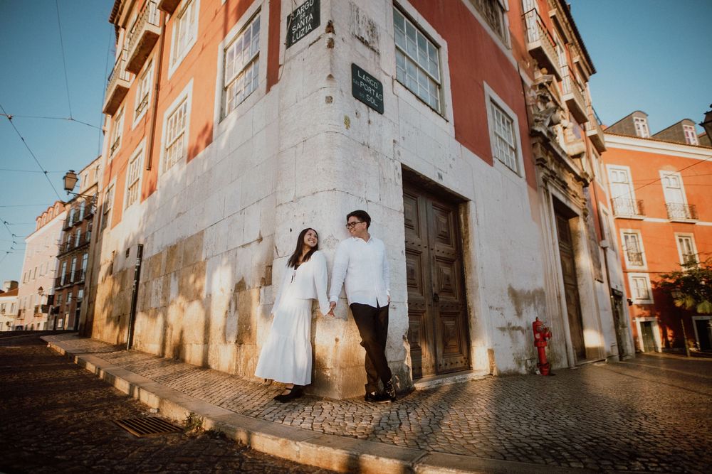A couple in white outfits stands at the corner of a rustic orange building on a cobblestone street in Lisbon, Portugal.