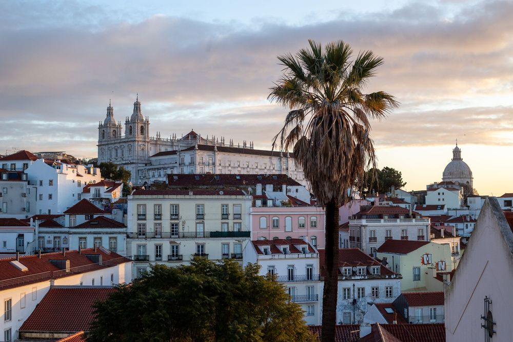 Panoramic cityscape view of Lisbon, Portugal at sunset with historic buildings and palm trees against a colorful sky.