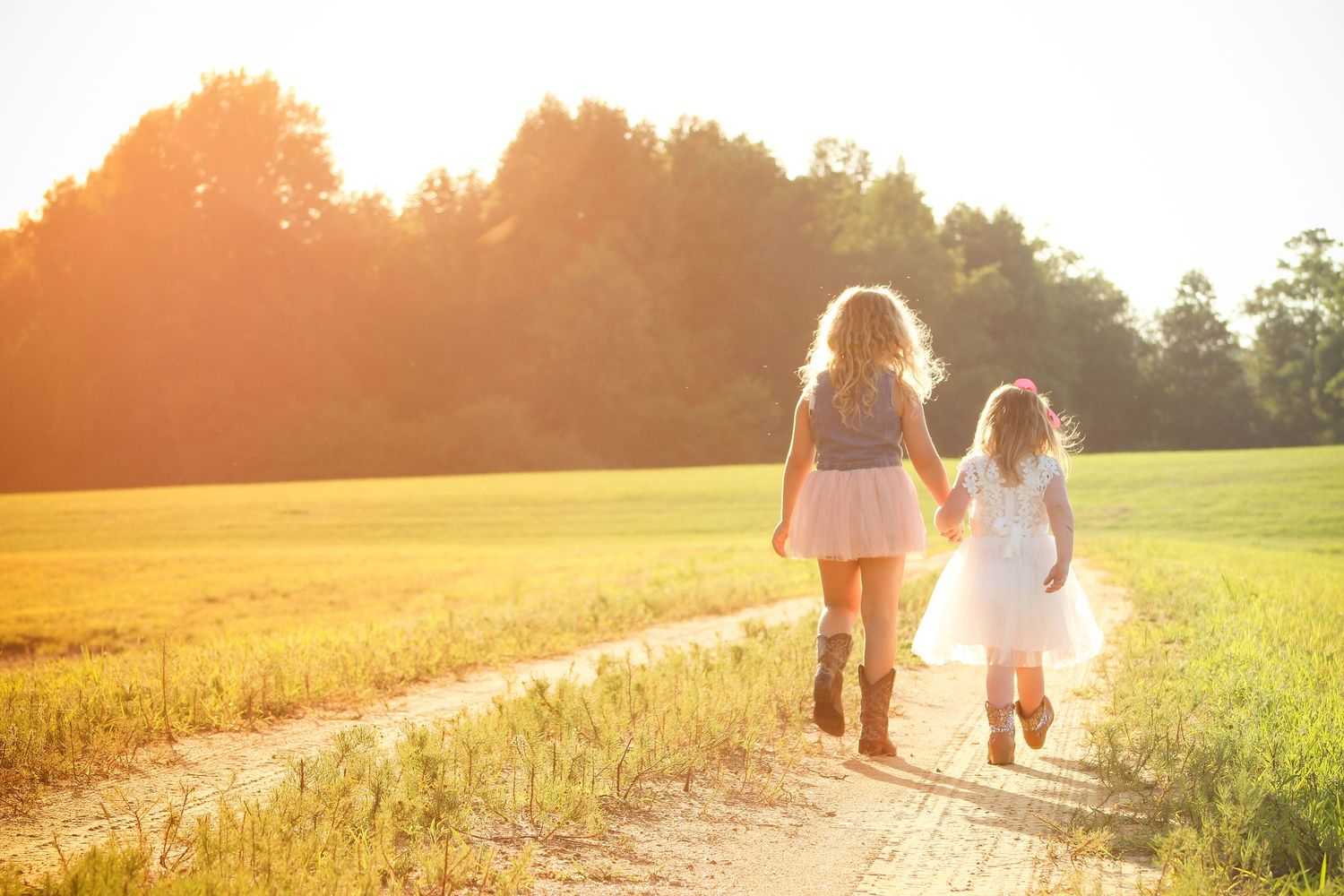 Two people walking hand in hand down a dirt path through a golden field at sunset with sunlight creating a warm glow.