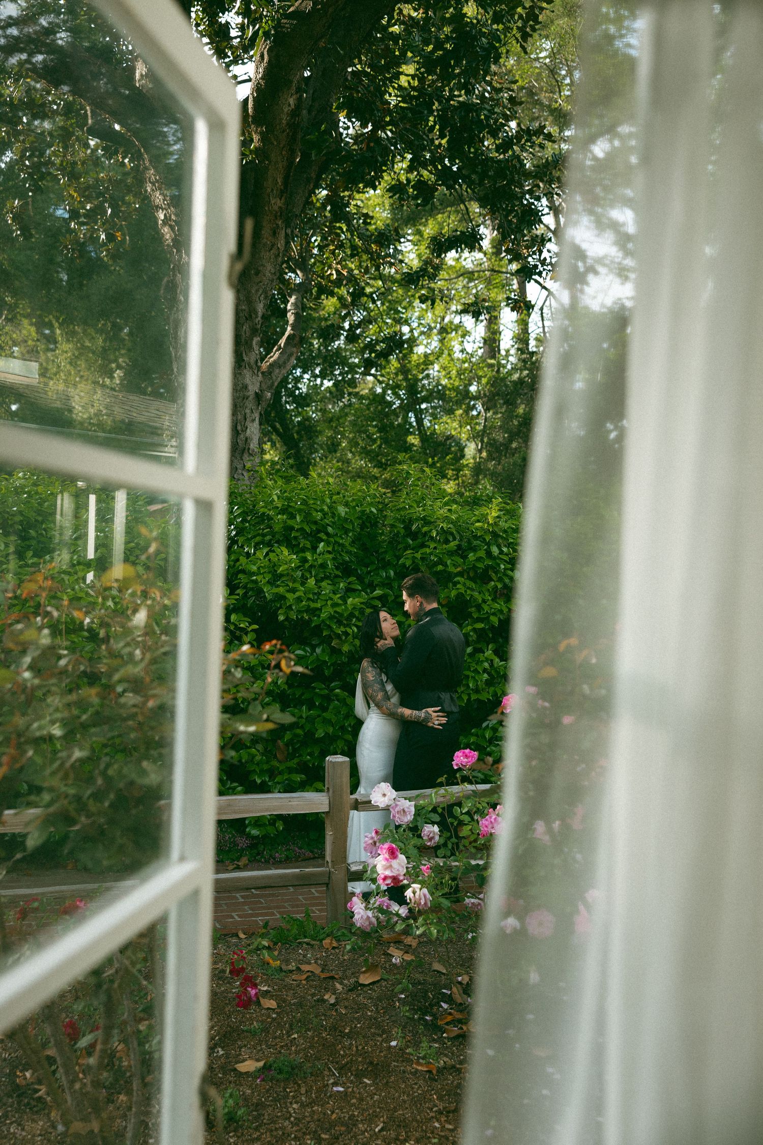 A couple poses together in a greenhouse with white columns and pink roses in bloom.