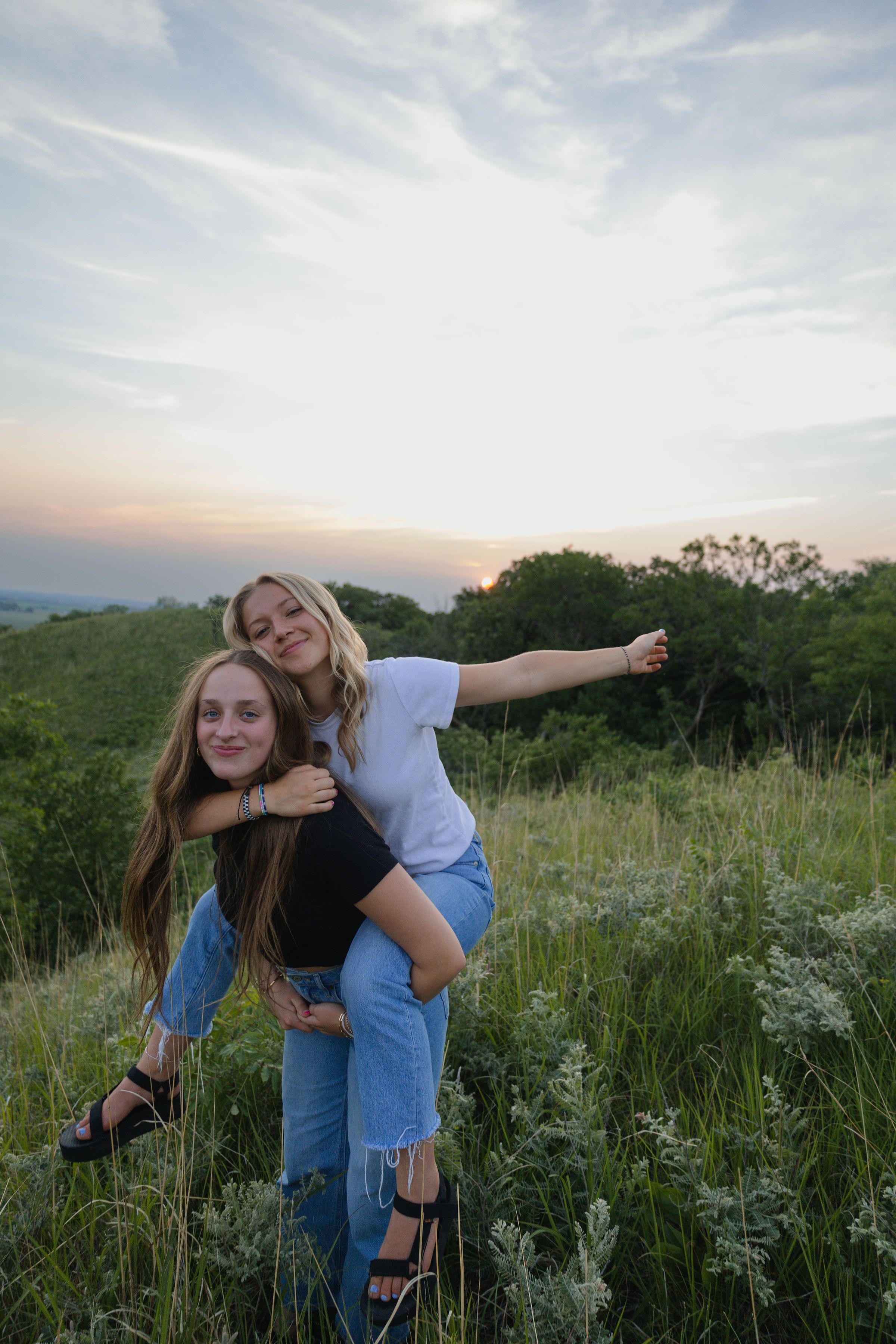 Sioux City, Iowa photographer. Two teenage girls pose at sunset in a green meadow, one giving the other a piggyback ride.