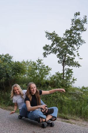 Sioux City, Iowa photographer. Two teenage girls laugh while riding a longboard skateboard down a road surrounded by greenery.