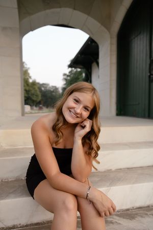 Sioux City, Iowa photographer. Smiling teenage girl in black strapless dress sitting on stone steps under arched entrance at dusk.