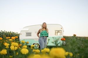 Sioux City, Iowa photographer. Young woman in green top stands in yellow flower field before vintage mint camper at sunset.
